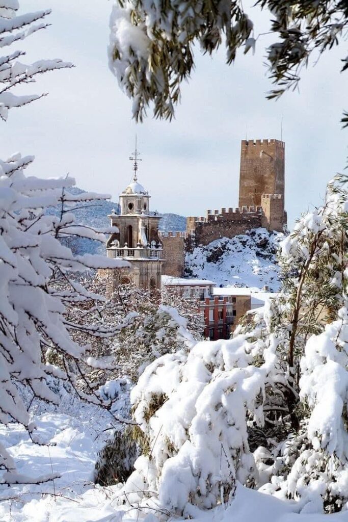 A snow-covered landscape reveals a medieval castle with a towering clock tower, nestled among snow-laden trees, as if protecting a slumbering princess within its wintery embrace.