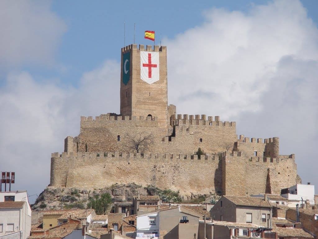 A medieval castle with high towers and fluttering flags is perched on a hill, unexpectedly overlooking a town with red-tiled roofs, as if awaiting the return of its princess.