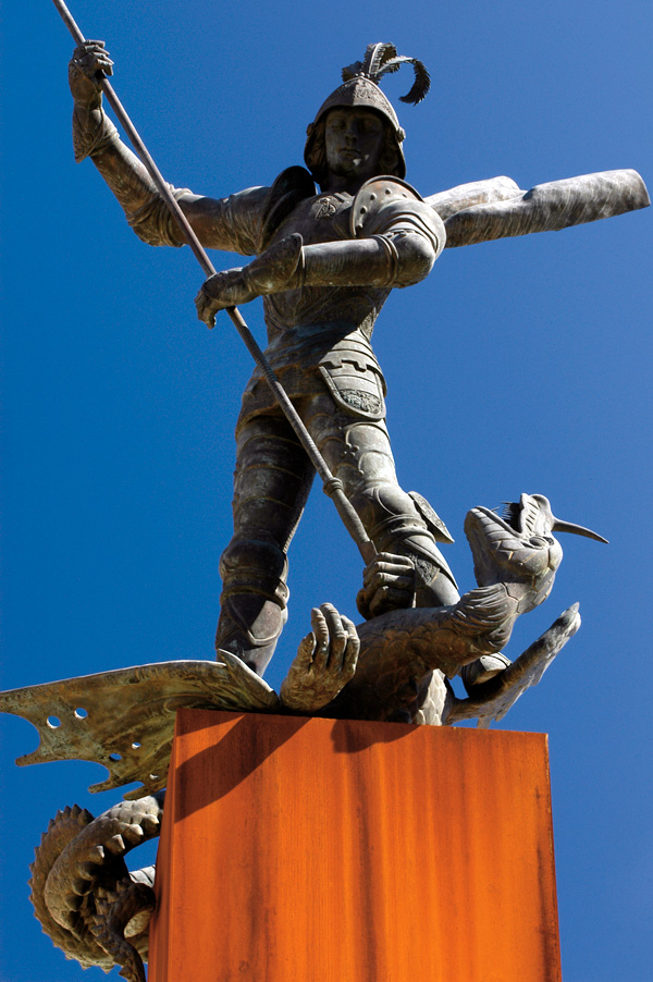A bronze statue of a knight in armor stands atop a dragon, holding a spear triumphantly against the backdrop of a clear blue sky, as if guarding the tower of an awaiting princess.