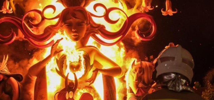 A fireman observes a large, decorative sculpture engulfed in flames during a nighttime event.