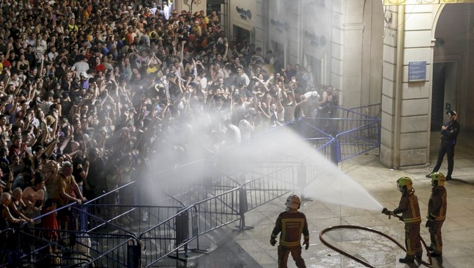 Naughty firefighters spray water to control a large crowd behind barricades outside a building at night.