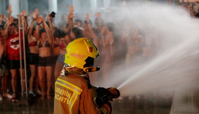 A naughty fireman playfully sprays water towards a crowd of people, some in swimwear, who appear enthusiastic behind the barrier.