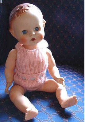 A vintage baby doll with curly hair, clad in a pink knitted outfit, sits dreamily on a blue patterned surface, evoking a sense of magic.