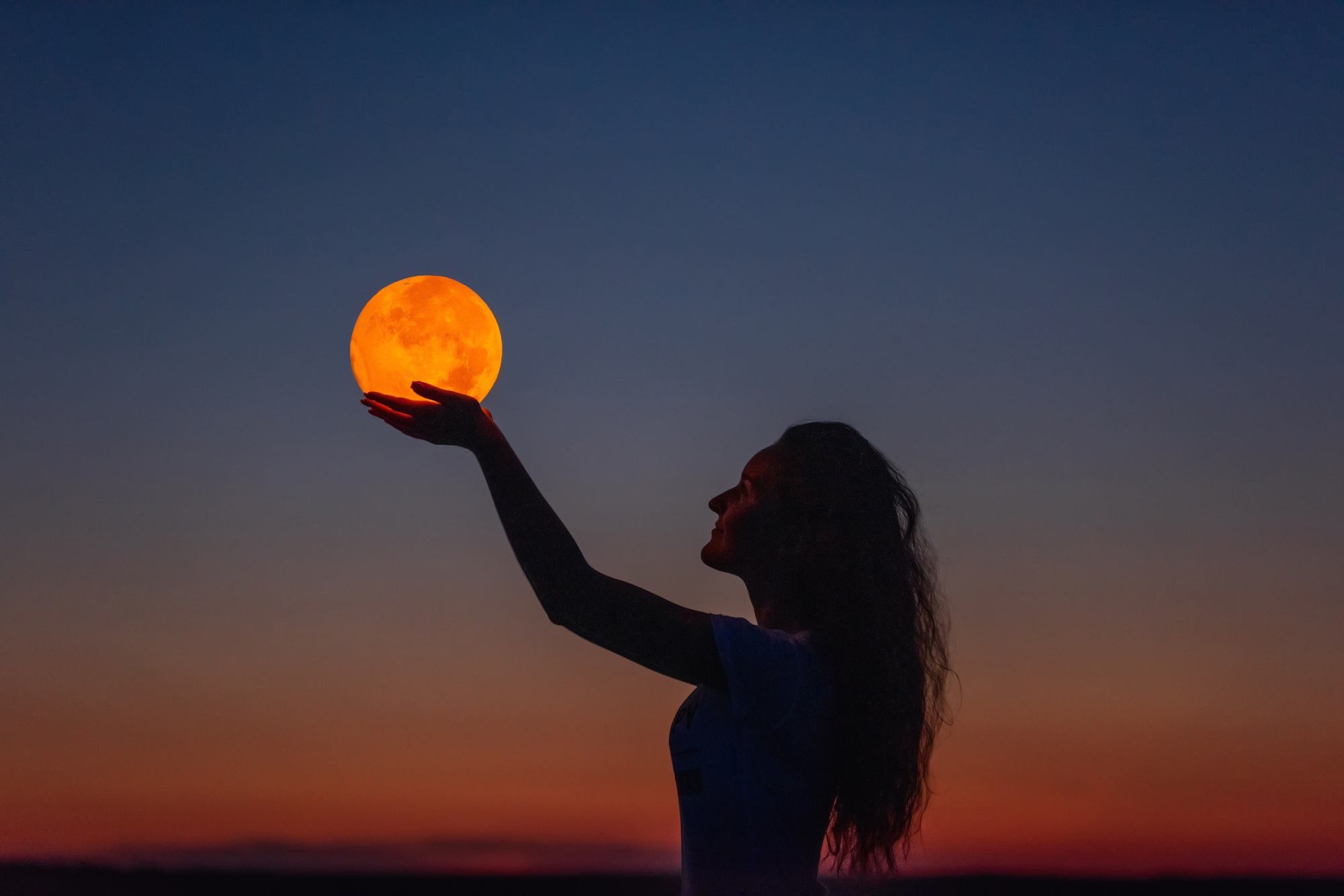 Silhouette of a woman holding her hands to the sky as if cradling a large, orange full moon at dusk.
