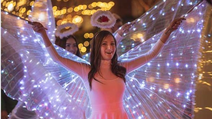 During a festive event in Alcoy, a woman dressed as an angel with glowing wings and a halo smiles brightly as the lights twinkle in the background, capturing the magical spirit that started this joyous celebration.