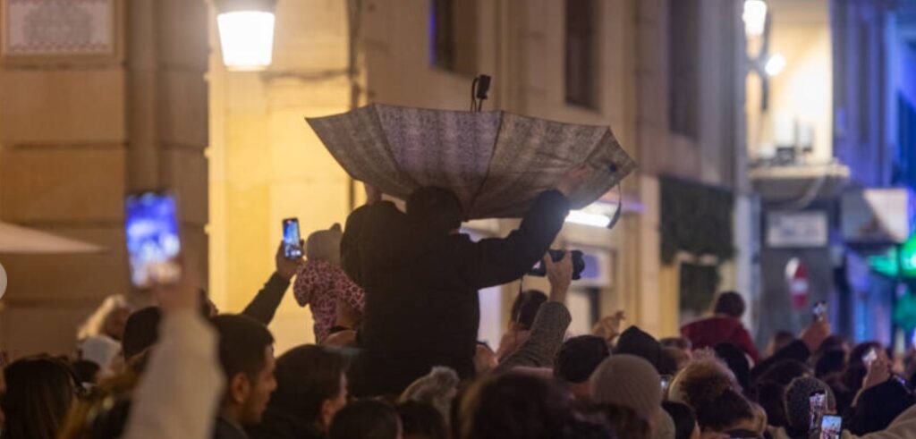 Amidst the bustling streets of Alcoy at night, a person holds an umbrella upside down with a mobile phone attached, capturing the scene where everything started.