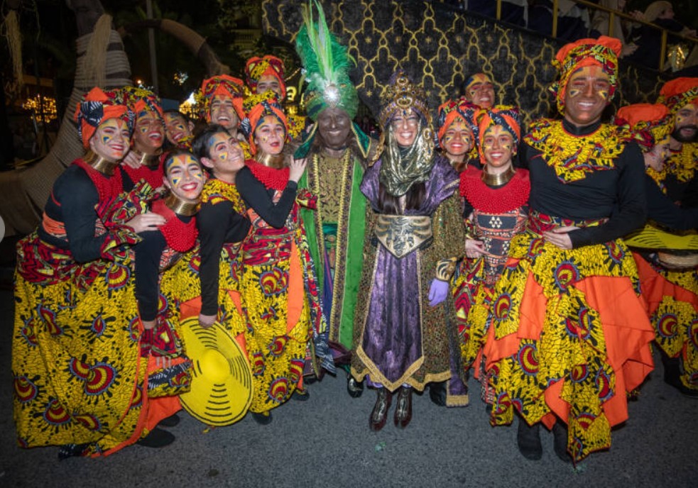 A group of people in vibrant, colorful costumes pose together, smiling at a festive event in Alcoy, where the tradition started, with intricate patterns and headdresses.
