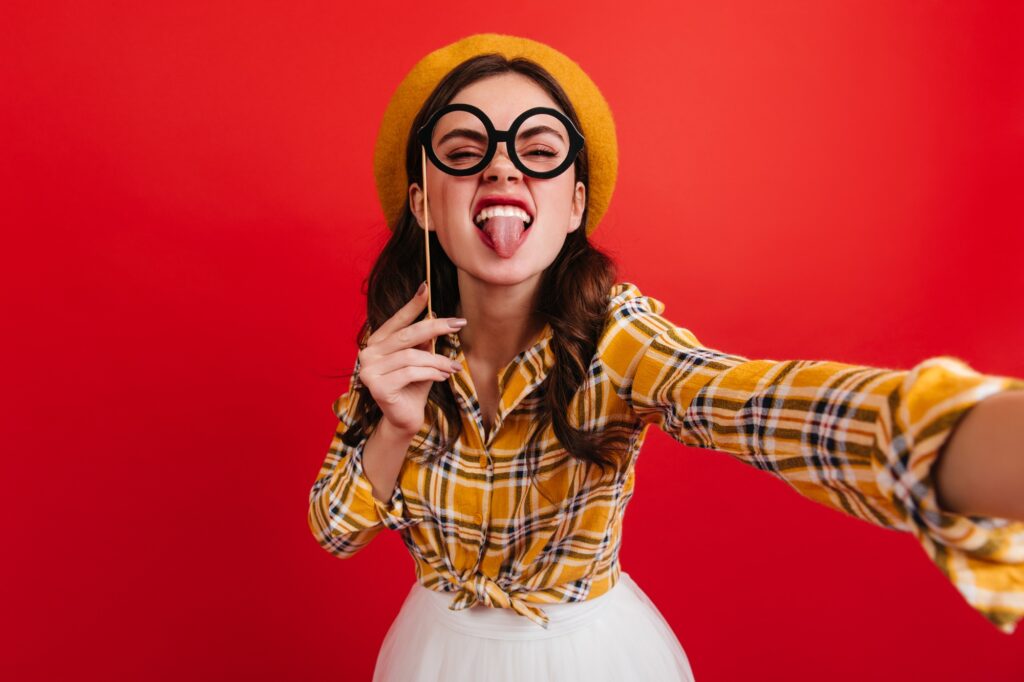Lady in beret and round glasses shows tongue. Selfie of naughty girl in bright outfit on red backgr