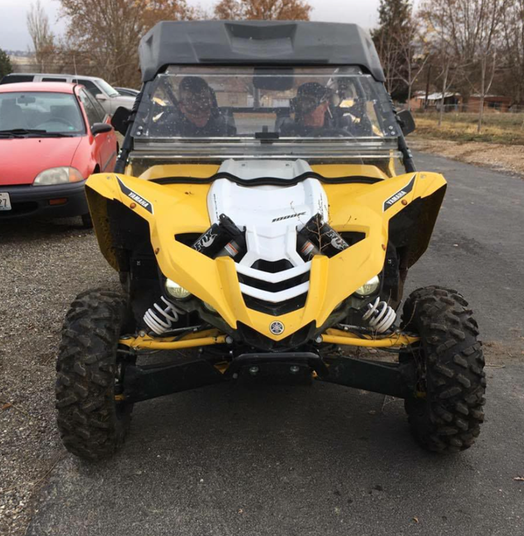A yellow off-road vehicle parked on a driveway, with two people inside. A red car is partially visible on the left. Trees and a house are in the background.