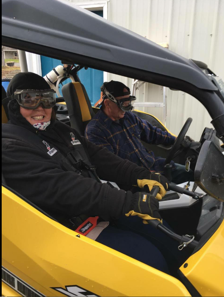 Two people wearing goggles in a yellow off-road vehicle; the passenger is smiling and the driver is focused ahead.