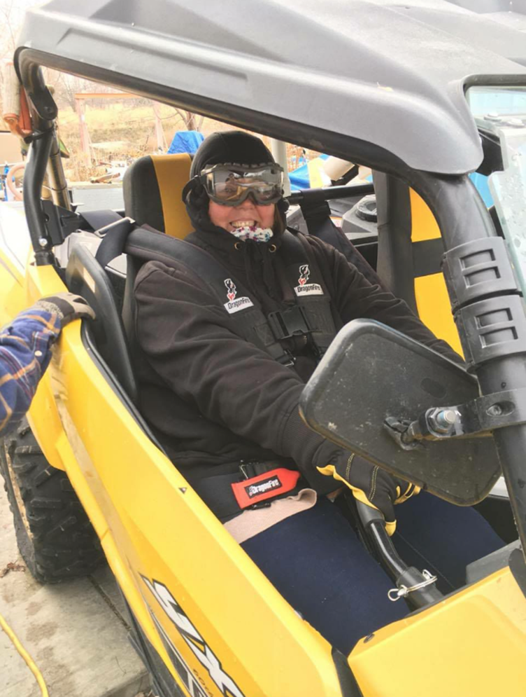 A person in winter gear sits in a yellow off-road vehicle, holding the steering wheel. The person is wearing goggles, a helmet, and a jacket.