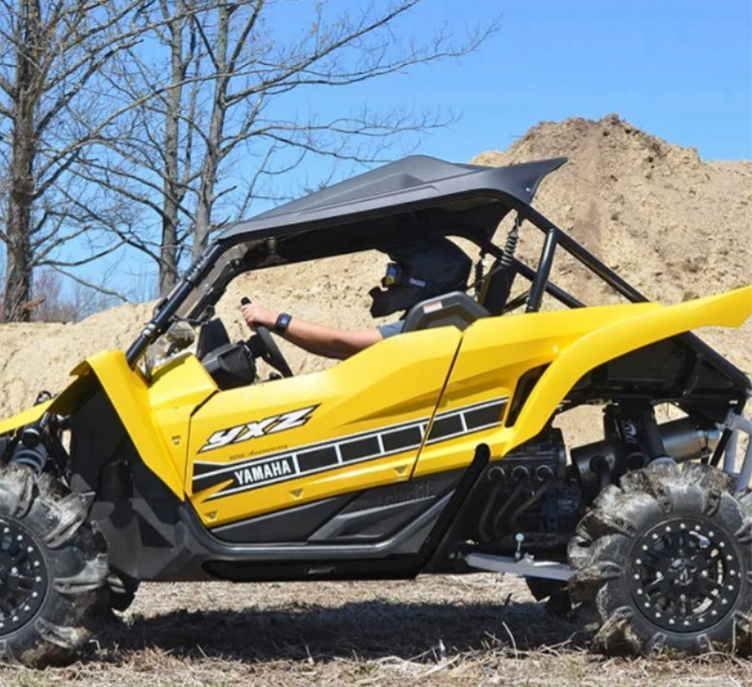 Yellow Yamaha YXZ side-by-side vehicle driving on a dirt path, with a person wearing a helmet and goggles. Trees and a dirt mound are visible in the background.