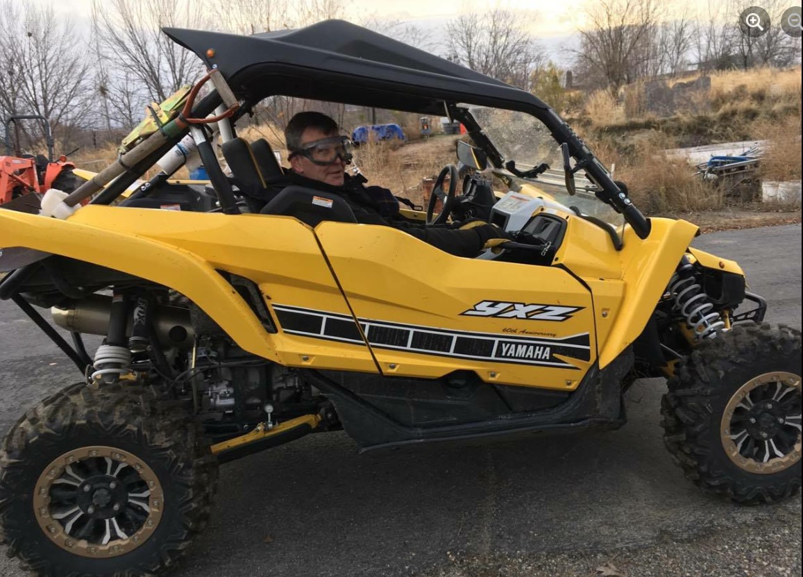 A person embarks on an adventure, driving a yellow Yamaha YXZ off-road vehicle equipped with a sturdy roll cage. With barren trees and grass in the background, this wild ride unfolds on a paved surface, promising thrills and excitement at every turn.