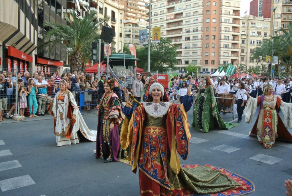 People in colorful, traditional costumes are participating in a parade on a city street brimming with spectators and buildings that will blow your mind.