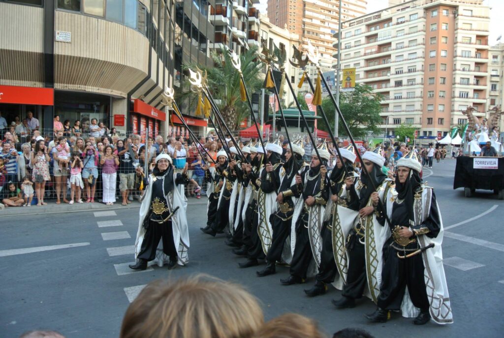A group of people in ornate black and white costumes march in a street parade, carrying long staffs. Spectators, ready to have their minds blown, line the street amidst tall buildings in the background.