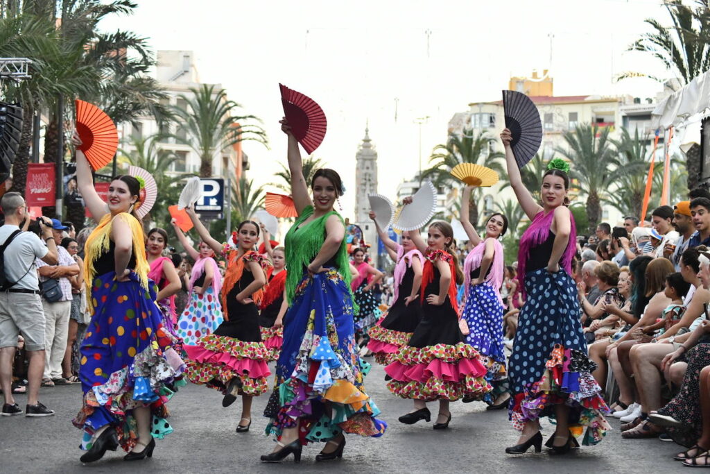 A group of dancers in colorful outfits performs with fans on the street, captivating a crowd. Palm trees and buildings set the backdrop for this stunning display that's sure to blow your mind.