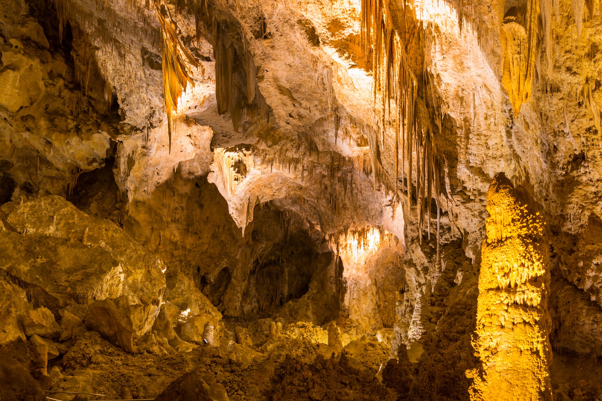 A cave interior with stalactites and stalagmites, illuminated by warm artificial lighting enhancing the rocky textures.