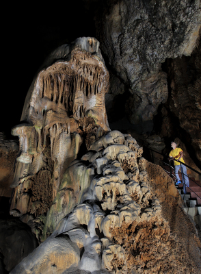 A person in a yellow jacket stands on stairs inside a cave, exploring hidden wonders as they observe large rock formations with stalactites and stalagmites.