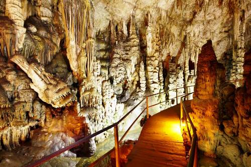 A wooden walkway with a railing passes through a cave filled with stalactites and stalagmites, illuminated by yellow lighting, offering a sense of surprise around every corner.
