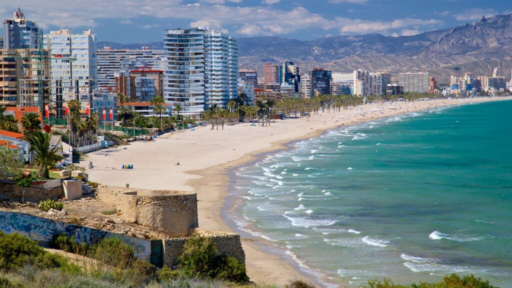 A coastal cityscape with tall buildings lining a sandy beach beside turquoise waves, where mountains rise in the background under a partly cloudy sky, offering a surprise view at every turn.