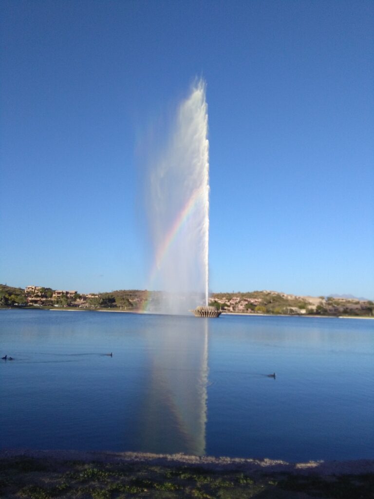 A tall water fountain of unexpected height shoots into the air from a lake, creating a visible rainbow in the spray under a clear blue sky.