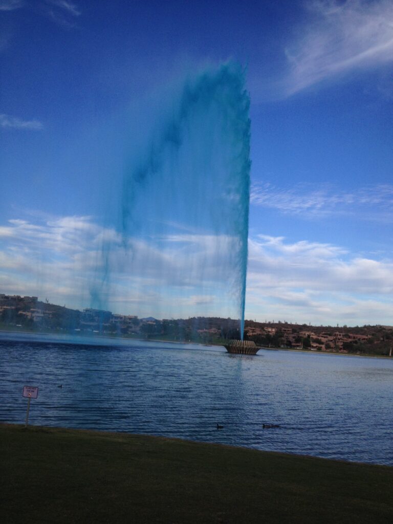 A tall fountain of unexpected height sprays a stream of bright blue water over a lake, with houses and a cloudy sky in the background.