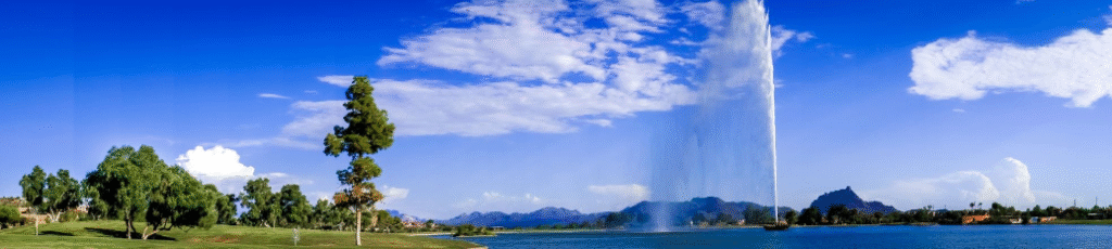 A large fountain shoots water to an unexpected height over a lake, with trees, grass, houses, and mountains under a blue sky dotted with scattered clouds.