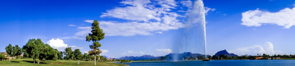 A large fountain shoots water to an unexpected height over a lake, with trees, grass, houses, and mountains under a blue sky dotted with scattered clouds.