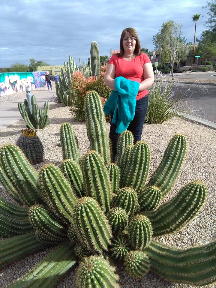 A woman stands outdoors on a gravel path behind a large cluster of cacti of unexpected height, holding a teal jacket, with more cacti and plants visible in the background.