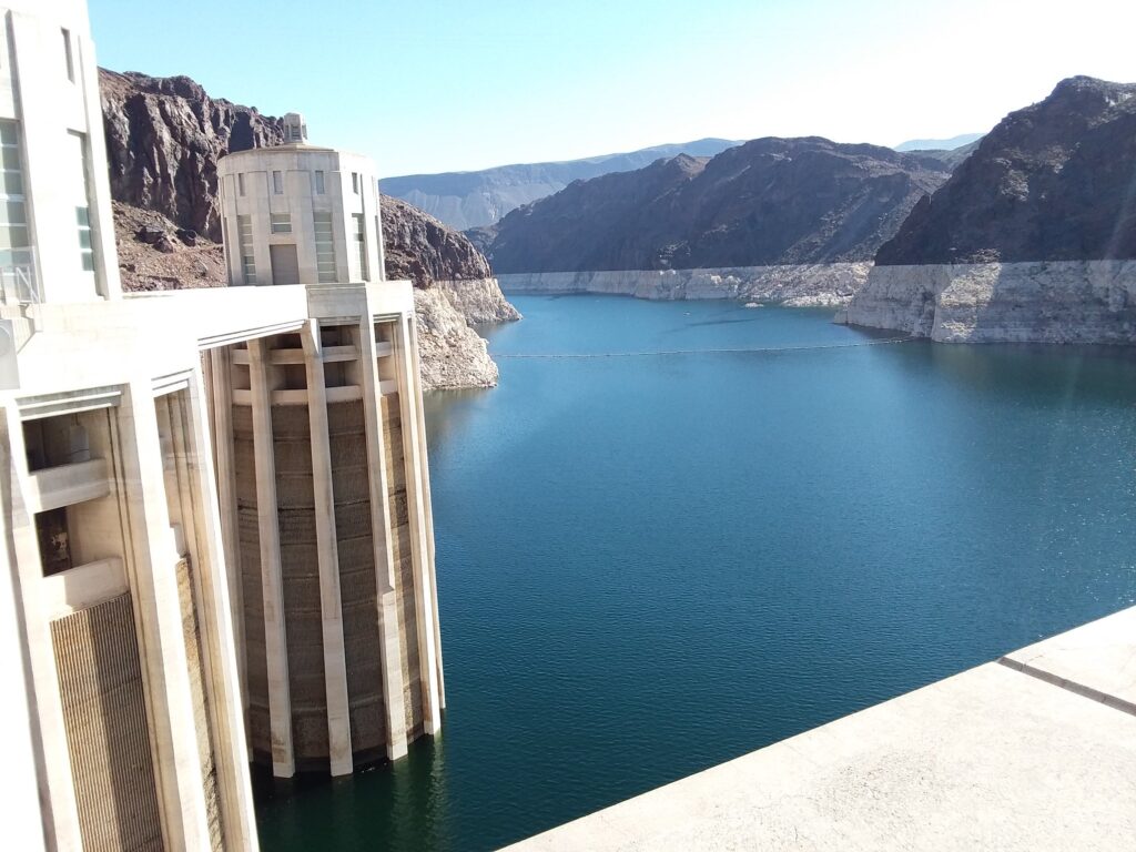 A large dam structure with intake towers stands beside a deep blue reservoir, nestled in the rocky, mountainous terrain—its massive concrete walls resembling the Belly of the Beast under a clear sky.