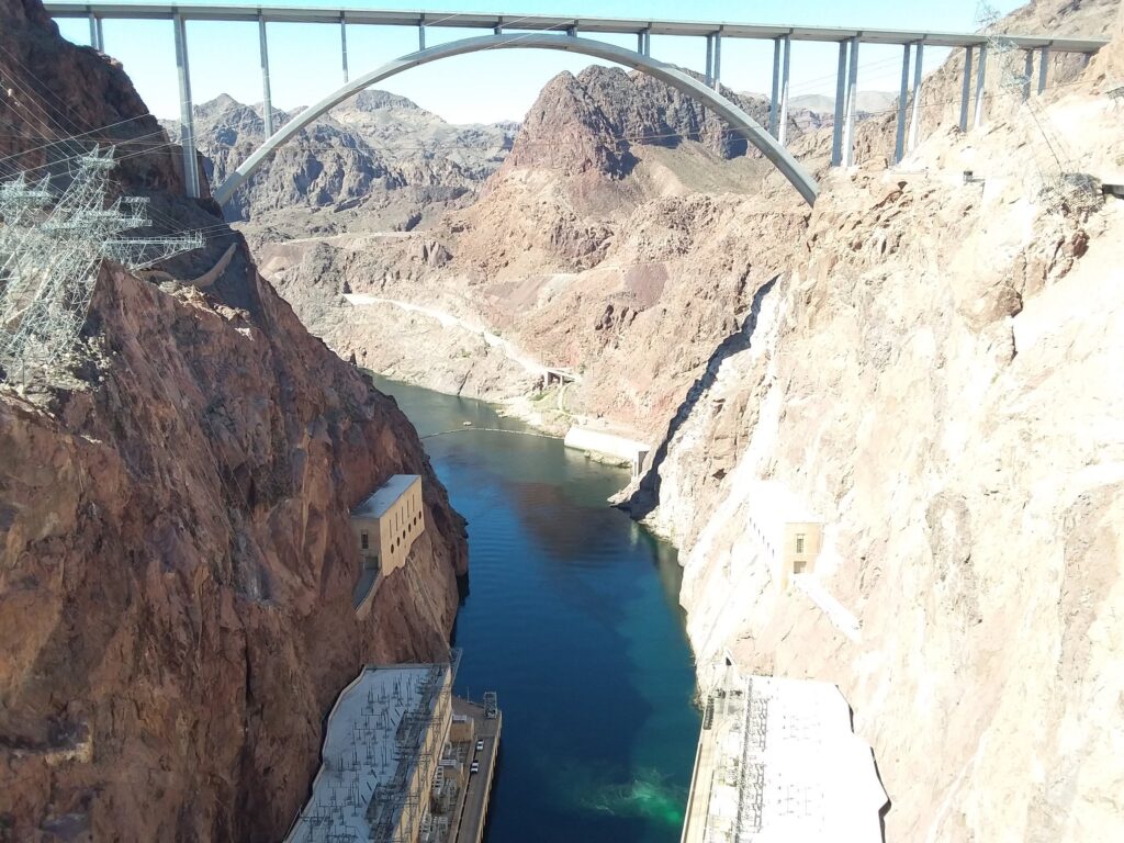 A large concrete arch bridge spans a deep canyon with steep rocky walls, crossing over a river and a dam below—plunging boldly into the belly of the beast.