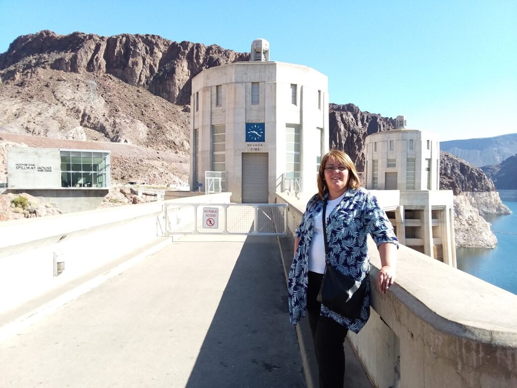 A woman stands on a walkway in front of a large dam structure—an entry Into the Belly of the Beast—with a clock, rocky hills, and water visible in the background under a clear sky.