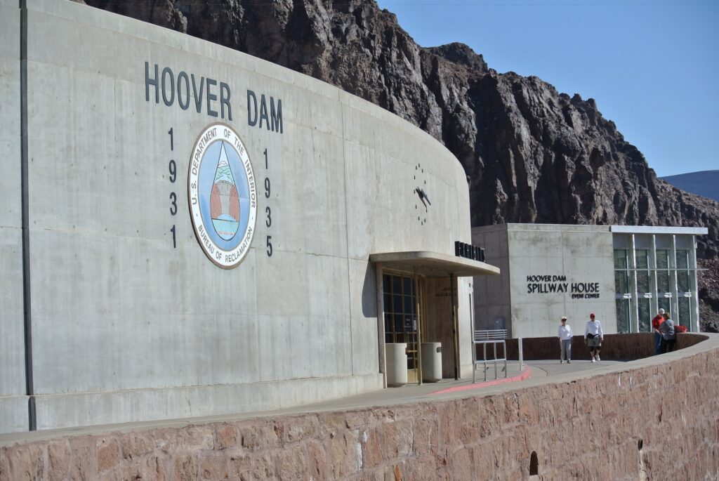 Hoover Dam exterior with the seal, construction dates 1931 and 1935, and the nearby Spillway House visitor center. Several people walk along the path, venturing Into the Belly of the Beast amid rocky cliffs in the background.