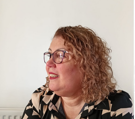 A woman with relatively curly, light brown hair and glasses looks to the left, wearing a patterned black and beige top, against a plain white background.
