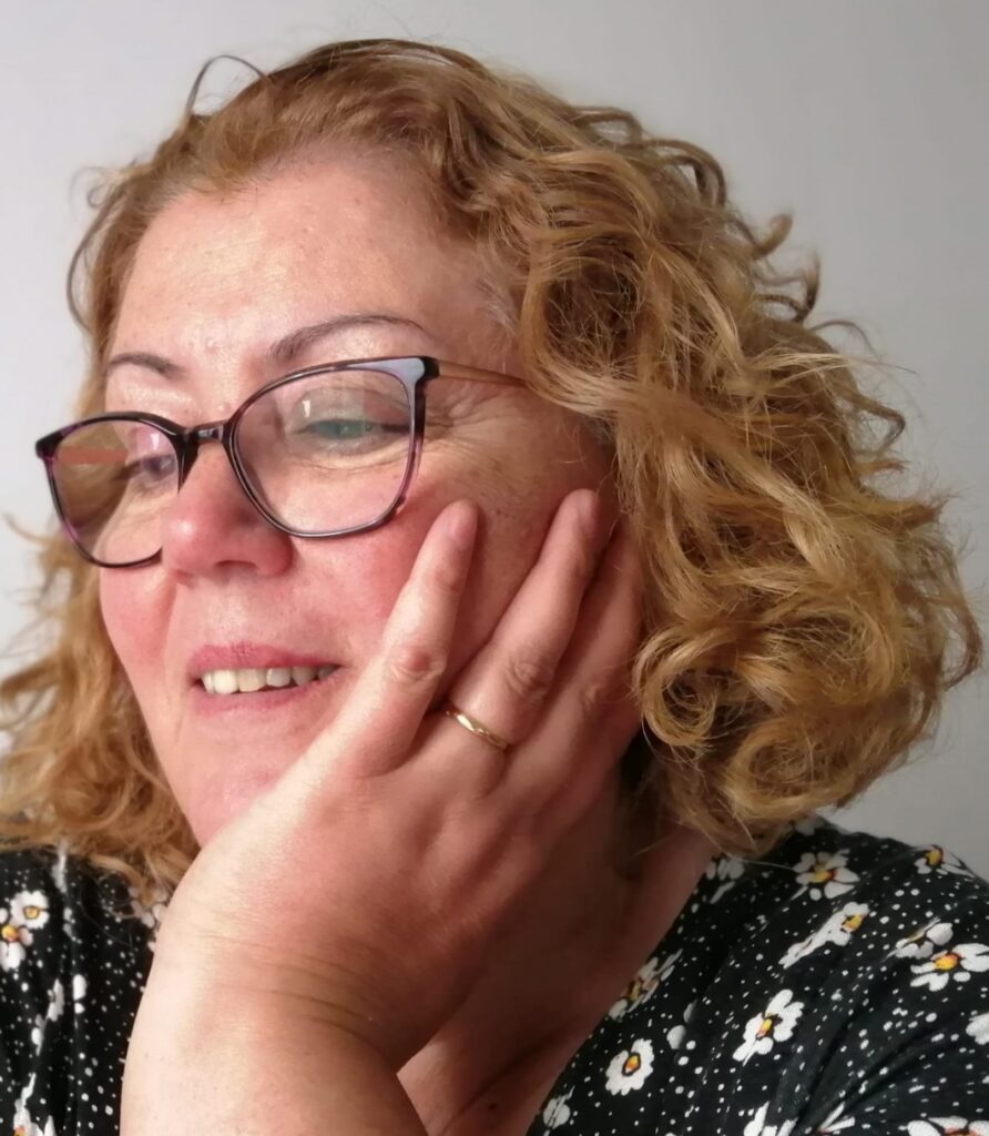 A woman with curly blonde hair and glasses rests her chin on her hand, wearing a floral top and a gold ring, appearing deep in thought about her self-worth against a plain background.