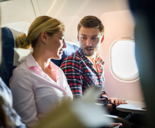 A man and a woman sit next to each other on an airplane, having a conversation by the window seat about self-worth and life beyond net worth.