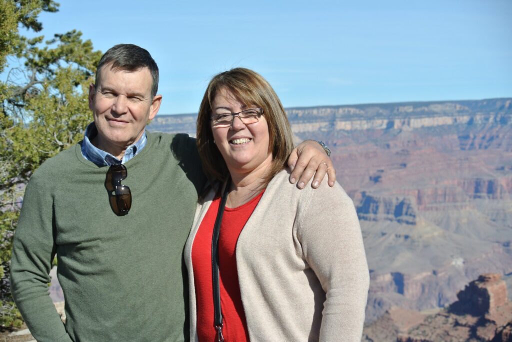 Two adults standing close together and smiling at the Grand Canyon, with layered rock formations in the background on a clear day—capturing the joy of travel and adventure.
