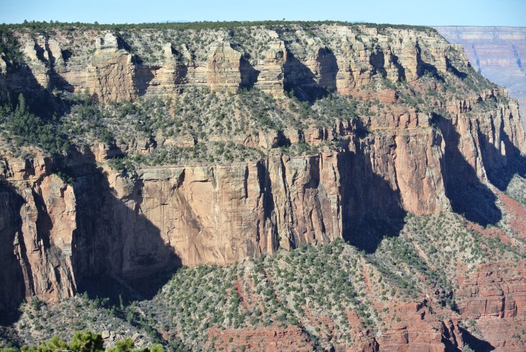 Steep rock cliffs with layers of red and tan stone, sparse vegetation, and deep shadows line the Grand Canyon wall under clear daylight—a perfect travel destination to explore by SUV.