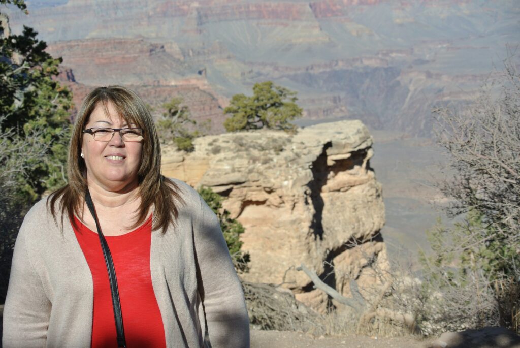 A woman with glasses and shoulder-length hair stands in front of a rocky cliff at the Grand Canyon, enjoying a sunny day of travel.