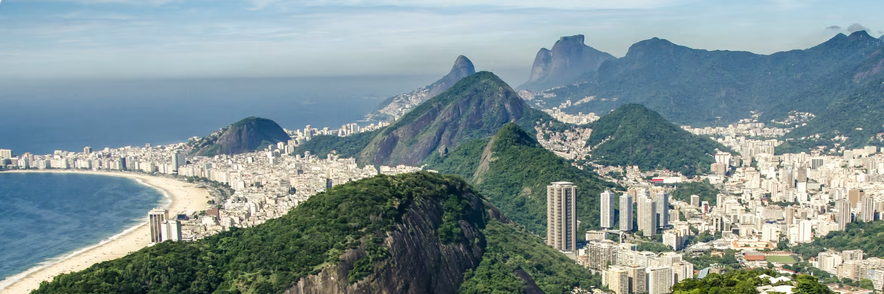 Aerial view of Rio de Janeiro featuring mountains, dense city buildings, and a long stretch of beach under a partly cloudy sky.