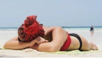 A person with red braided hair lies face down on a towel at a sandy beach, wearing a red and black swimsuit. The ocean and other beachgoers—perhaps classmates on a class trip—are visible in the background.