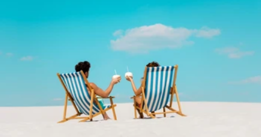 Two people in swimsuits sit in blue and white striped beach chairs on sand, facing the sky, holding drinks with straws under a clear blue sky.