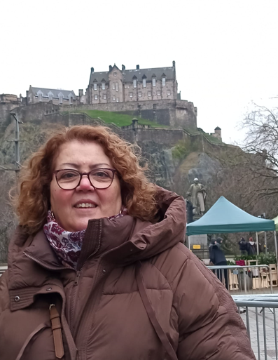 A woman in a brown coat and scarf stands outdoors with Edinburgh Castle visible in the background, where echoes of Burke and Hare linger in the historic atmosphere.