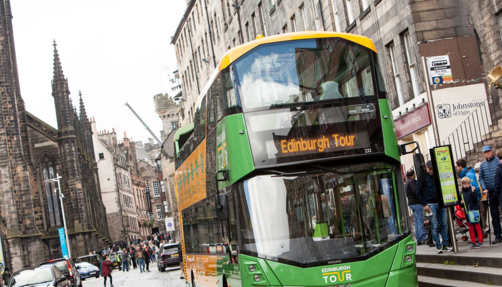 A green double-decker Edinburgh Tour bus drives along a cobbled street lined with historic stone buildings and pedestrians, echoing the infamous history of Burke and Hare in the shadowy corners of the city.