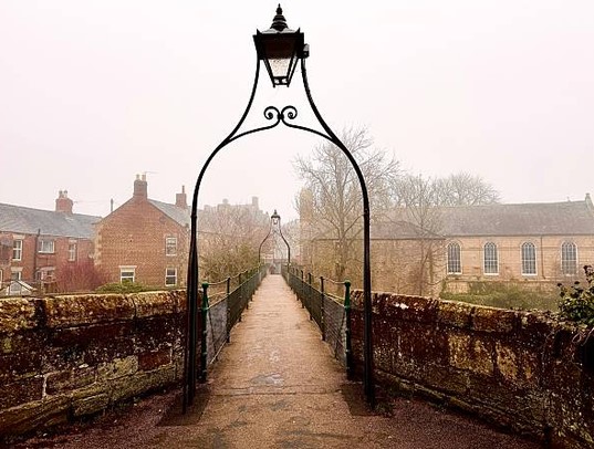 A stone footbridge crosses the brook, with wrought iron lamp-posts leading towards brick buildings and a church in Morpeth on a foggy day.