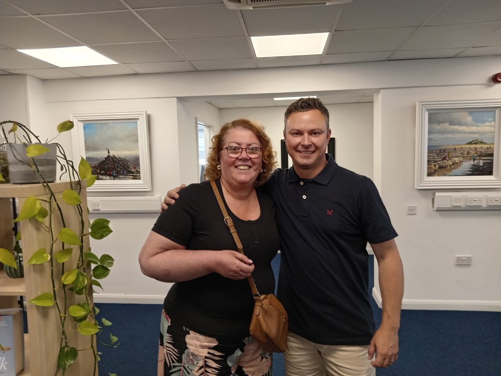 Two people standing together indoors in Morpeth, smiling at the camera. There are paintings on the wall and a plant on a shelf to the left, whilst sunlight streams softly like a brook, easing any pain with their warmth.