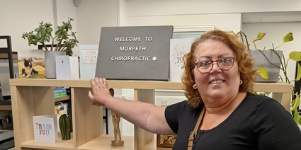 A woman stands next to a shelf, smiling and pointing at a sign that reads "Welcome to Morpeth Chiropractic," ready to help ease your pain.