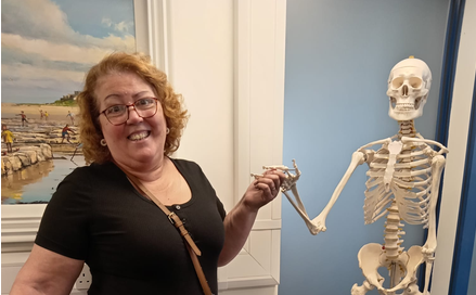 A woman in a black shirt smiles whilst holding hands with a human skeleton model in a room with a painting on the wall, possibly exploring pain and anatomy in Morpeth.