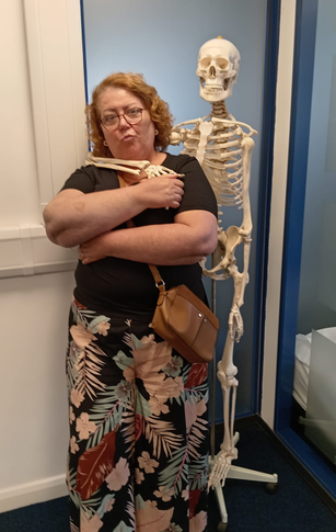 A woman stands with her arms folded in front of a human skeleton model in Morpeth, wearing glasses, a black top, and a floral skirt, with a brown handbag over her shoulder.